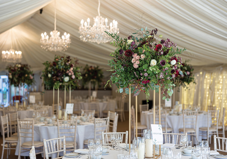 Statement floral centrepiece on gold stand styled on wedding reception tables inside a marquee