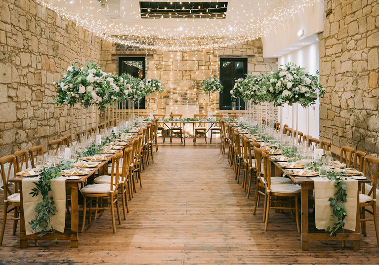 Stone-walled wedding reception with long wooden tables, white flowers and fairy lights planned by Pebblefish Occasions & Events.