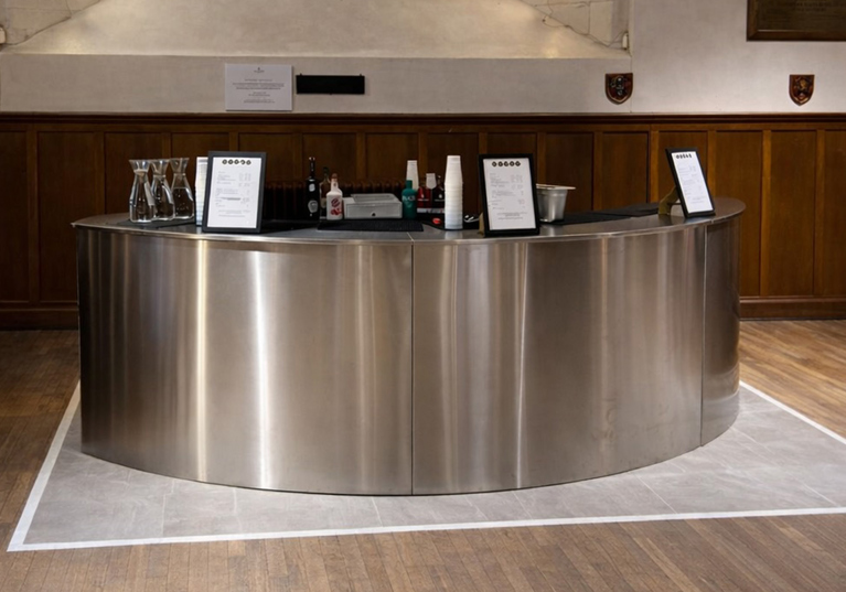 Curved stainless steel mobile bar by Sips Events positioned inside a historic event venue for drinks service.
