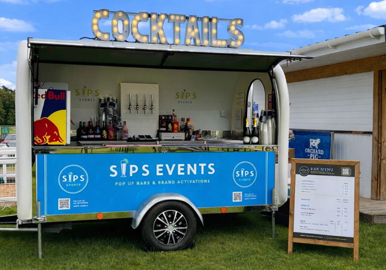 Mobile cocktail bar trailer by Sips Events set up outdoors with illuminated cocktail signage and drinks service station.