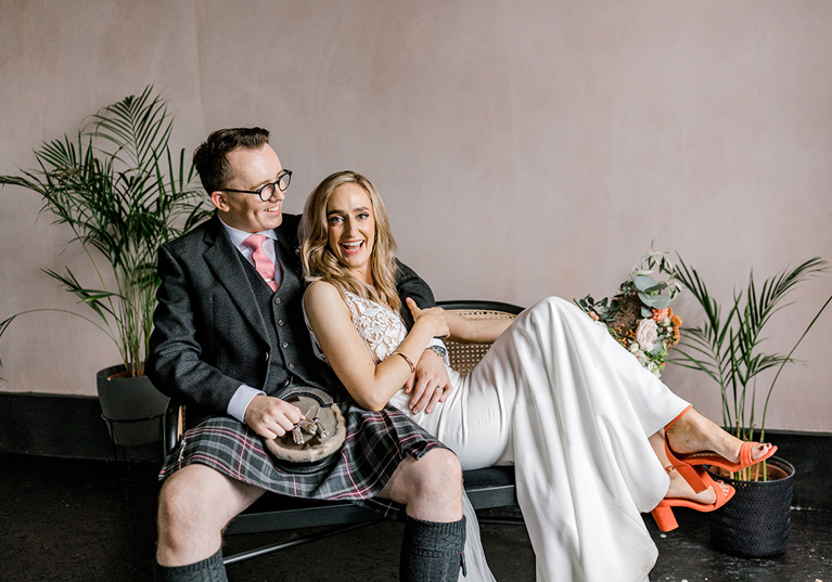 Bride and groom laughing together on a rattan bench, with the groom wearing a kilt and the bride in a white wedding outfit with orange heels.