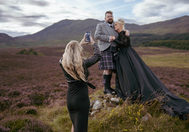 Louise, owner of Millar Moments, in action, capturing a dramatic image of a bride in a black wedding dress and her groom with Scottish hills as their backdrop.