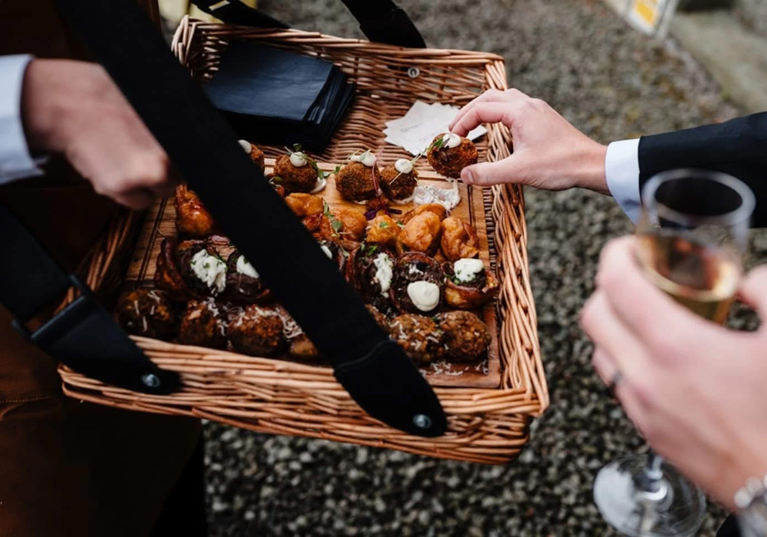 Tatties & Tipples team member holds wicker basket filled with canapés as guest holding glass of Champagne reaches for one.