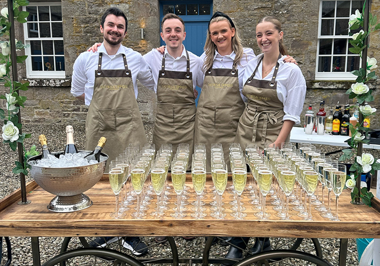 Four members of the Tatties & Tipples Event Catering team, standing in front of a cart filled with glasses of Champagne.