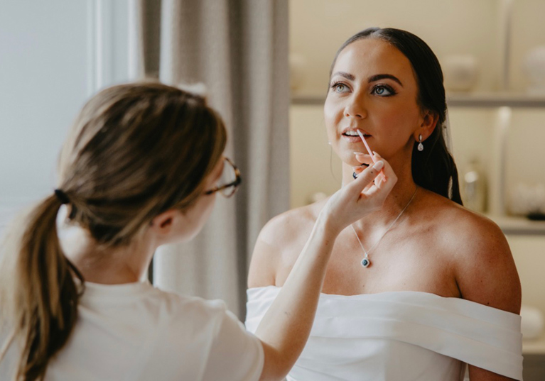 Wedding morning moment as Hannah Inglis applies lip gloss to a bride wearing an off-the-shoulder wedding dress