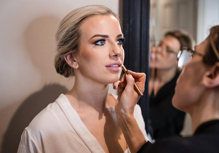 Bridal makeup artist applying lip liner to a bride in front of a mirror during wedding preparations