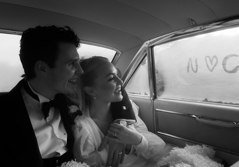 Black and white photo of newly married couple laughing together inside their wedding car with “N loves C” written on the window