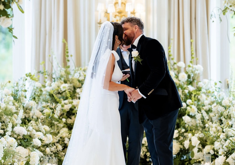 Bride and groom sharing a kiss during their wedding ceremony at Turnberry, framed by large-scale white floral installations and soft candlelight in an elegant Scottish venue.