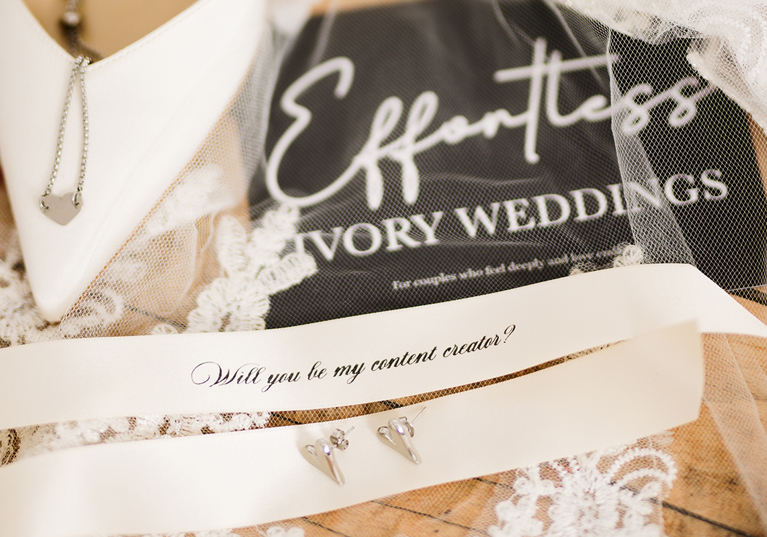 Bridal detail flat lay featuring jewellery, lace wedding accessories and a personalised “Will you be my content creator?” ribbon styled by Effortless Ivory Weddings