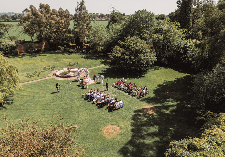 Aerial view of an outdoor wedding ceremony taking place on a large lawn surrounded by mature trees.