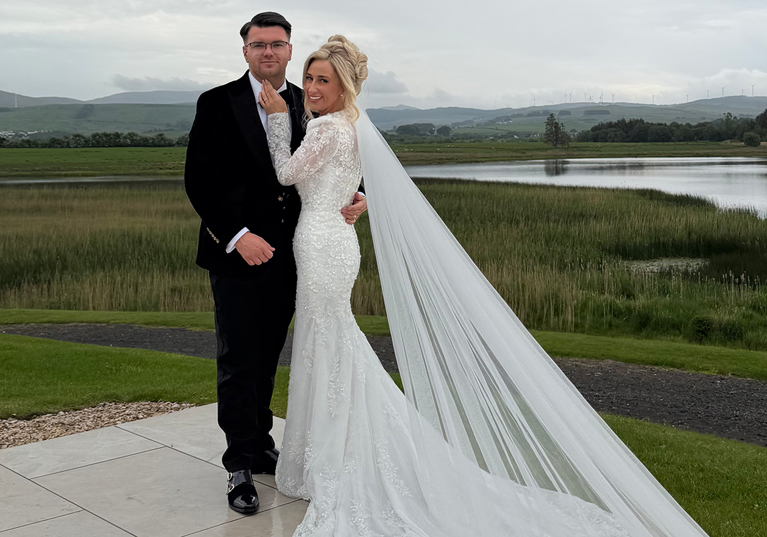 Bride and groom pose outside beside a scenic loch, with the bride’s long veil trailing across the terrace.
