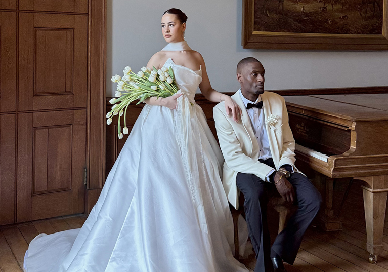 Bride holding white tulips stands beside the groom in a cream tuxedo jacket next to a grand piano.