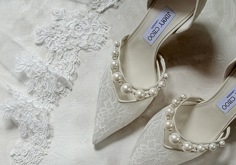 White lace Jimmy Choo bridal shoes decorated with pearls displayed beside an embroidered wedding veil.
