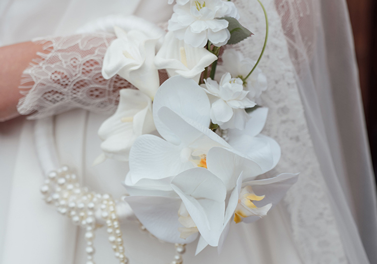 Close-up of white orchid and calla lily bridal flowers styled with pearl strands against a lace wedding dress.