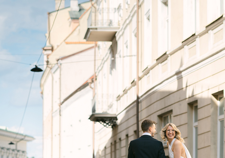 Candid wedding photograph by Goda and Justas Photography of newlyweds walking hand in hand along a city street, with the bride laughing in a flowing white halterneck dress.