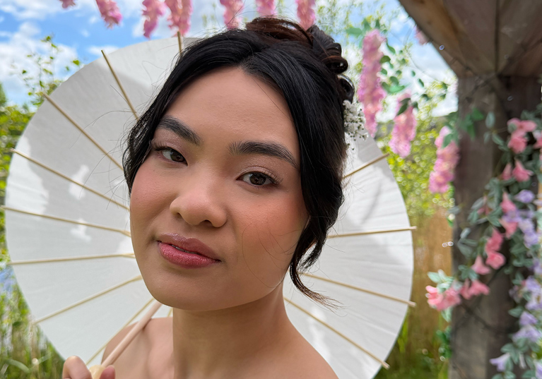 Bride holding a white parasol in a garden, showing soft pink bridal makeup and an elegant updo by Bridal by Regina.