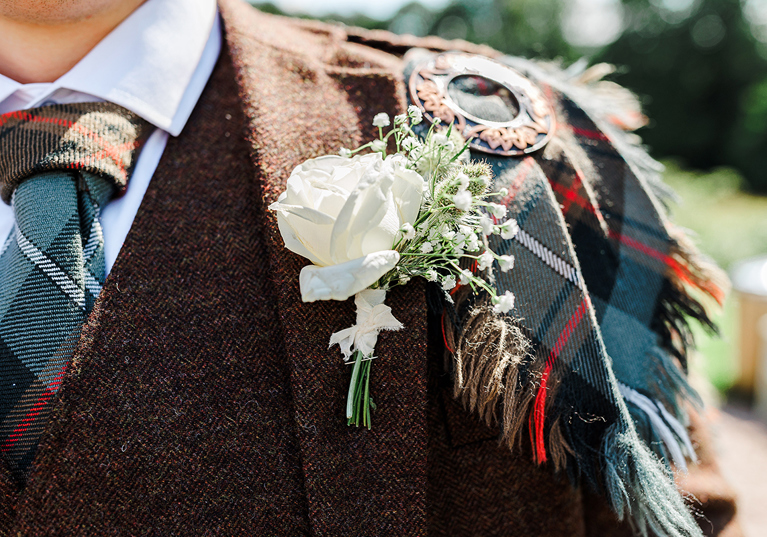 Close-up detail of groom’s Scottish wedding kilt, tartan sash and white floral buttonhole.