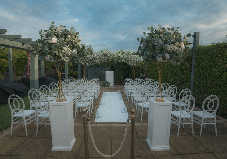 Outdoor ceremony set up with white chairs, white columns at top of aisle and floral installations.