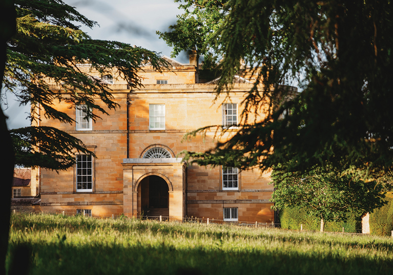 External view of Newton Don House from behind trees with tree shadows and sun shining on grass in foreground.