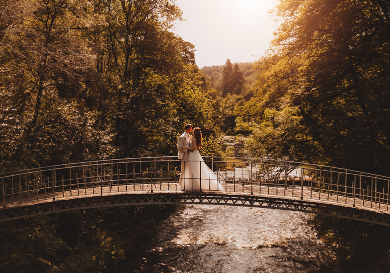Bride and groom standing on a bridge above the river at Broomholm Manor, a luxury exclusive-use wedding venue in Dumfriesshire.