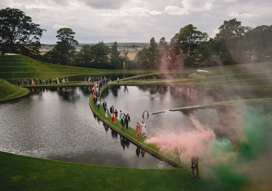 Wedding guests walking beside the sculpted ponds at Jupiter Artland with colourful smoke filling the grounds
