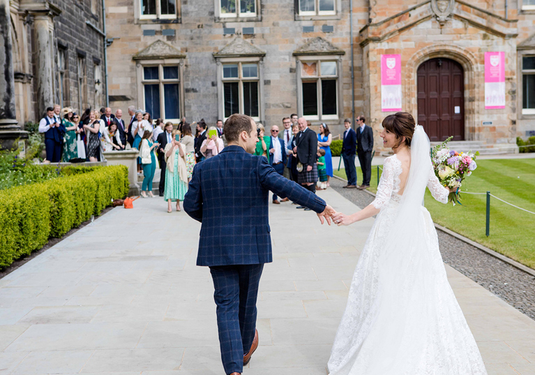Bride in a lace wedding dress and groom in a blue checked suit walk hand in hand outside the historic University of St Andrews buildings as guests watch.