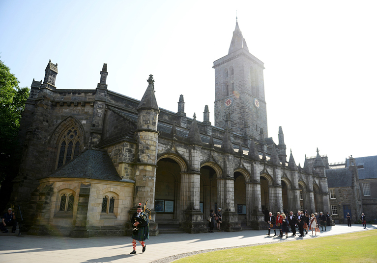 Traditional Scottish bagpiper leads a wedding party past the historic St Salvator's Chapel at the University of St Andrews.