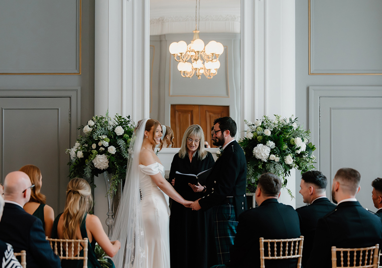 Bride and groom during their wedding ceremony at The Edinburgh Grand, framed by white and green floral arrangements.