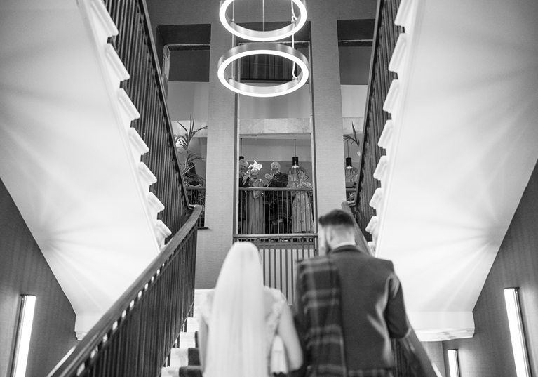 Black and white image of a bride and groom walking upstairs at The Clayton Hotel in Glasgow city centre, while guests look on and clap at the top.