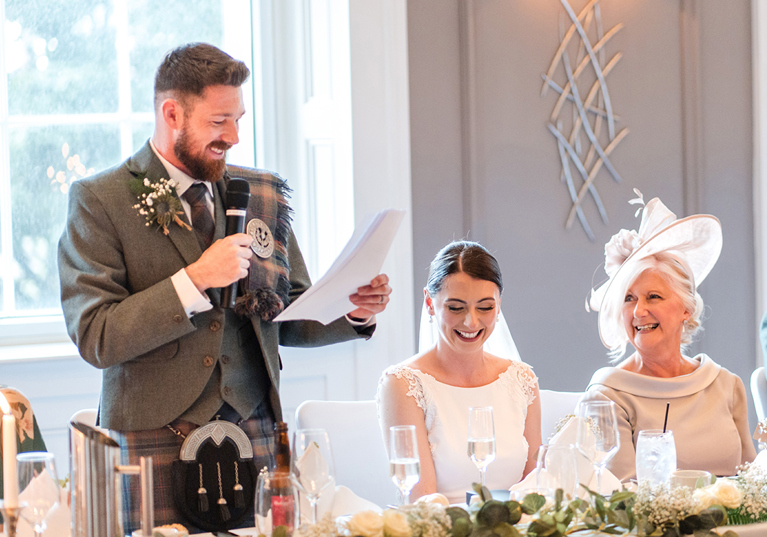 Groom makes speech at top table while bride and mother of the bride laugh at their wedding breakfast at The Clayton Hotel in Glasgow city centre