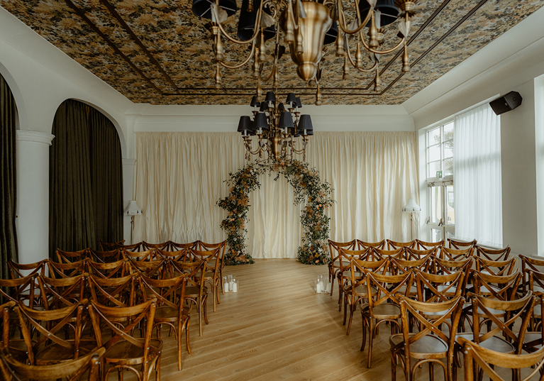 Indoor wedding ceremony setup at Dornoch Station Hotel with wooden cross-back chairs, floral arch and chandelier ceiling