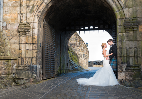 Bride and groom standing beneath the historic stone archway entrance at Edinburgh Castle during a romantic Scottish castle wedding photoshoot.