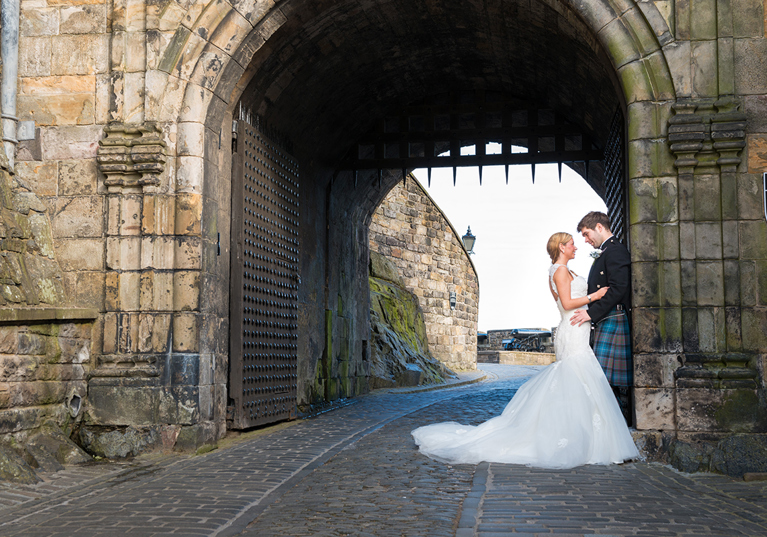 Bride and groom standing beneath the historic stone archway entrance at Edinburgh Castle during a romantic Scottish castle wedding photoshoot.