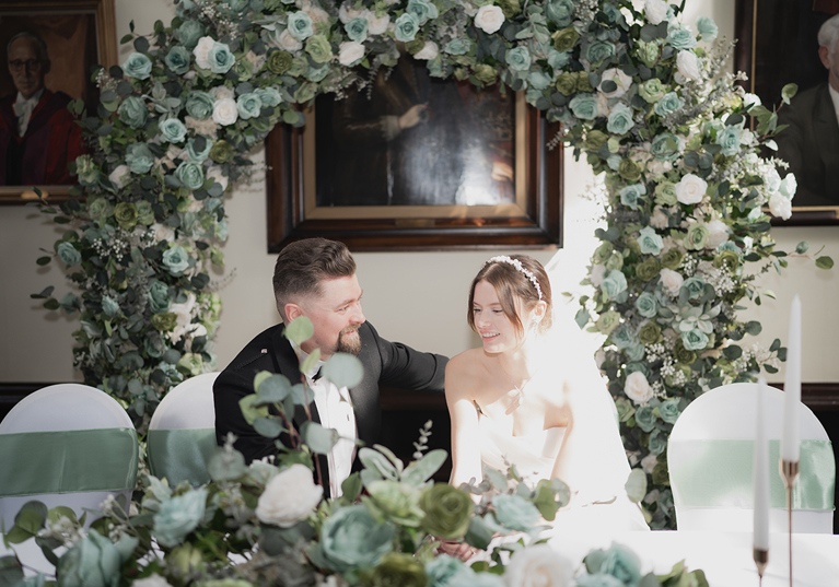 Bride and groom sit at top table in front of floral hoop.