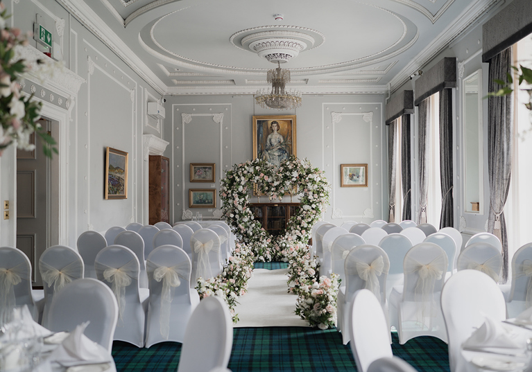 Ceremony set up for photoshoot at 1599 at the Royal College in Glasgow, with white chair covers and cream chiffon bows, and pink and white flowers lining the aisle to a large love heart shaped floral arch at the altar.