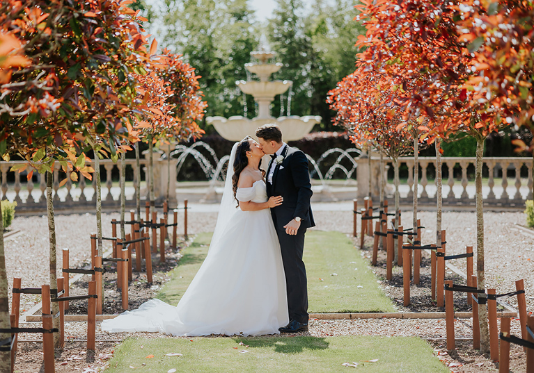 Newlywed couple kissing beside fountain and landscaped gardens at Ingliston Estate and Country Club wedding venue