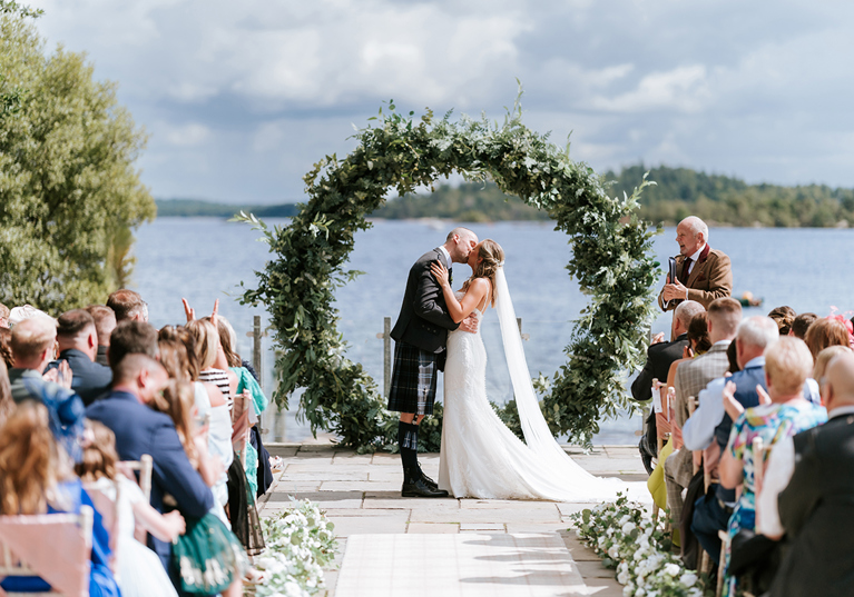 Bride and groom kiss at altar in waterside wedding ceremony with green hoop arch in background.