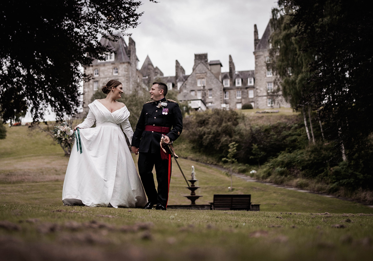 Bride and groom walking hand in hand in the gardens of Atholl Palace Hotel, with the historic Scottish castle-style hotel in the background; bride in a classic off-the-shoulder gown and groom in military dress uniform.