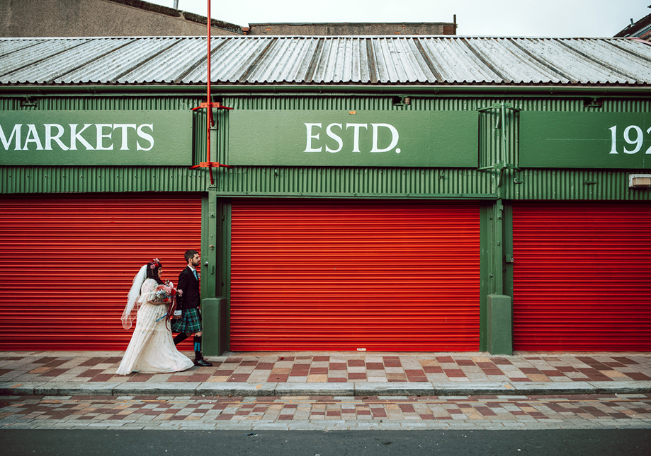 Bride and groom walk through Barra's Market in Glasgow, with red shutters behind them and "Markets Estd 1921" above them.