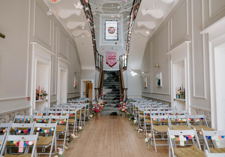 Wedding ceremony setup in the grand hall at Netherbyres House with aisle seating, staircase backdrop and rainbow pom-pom decor