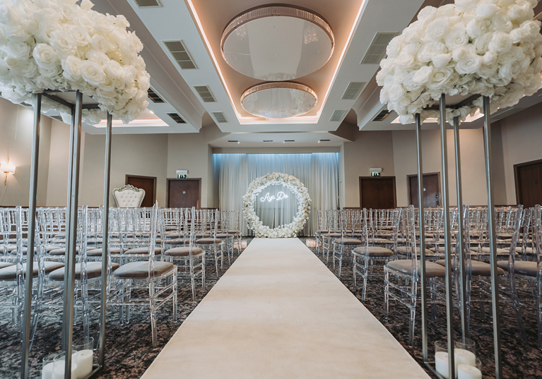 Wedding ceremony set up with clear chairs, white flowers on silver stands and a white flower hoop at altar with "Aye Do" lights