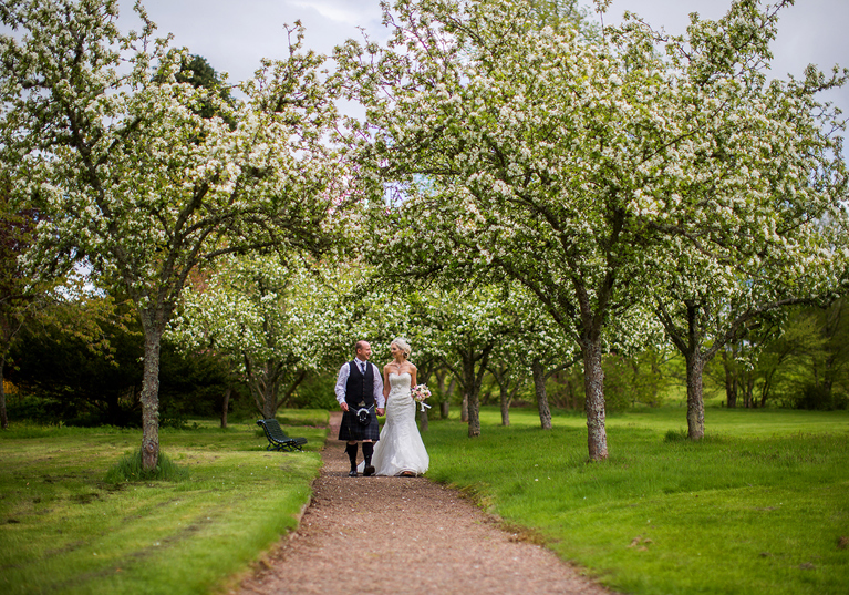 Bride and groom walk tree-lined path on the grounds of SCHLOSS Roxburghe in Kelso, captured by Parris Photography.
