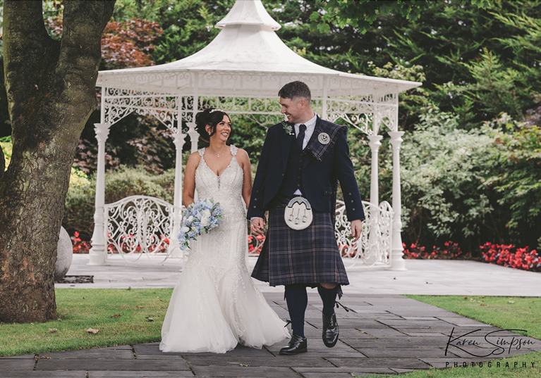 Bride and groom walk through gardens at Carlton Hotel in Prestwick, they are looking at each other and the bride is holding a baby blue bouquet