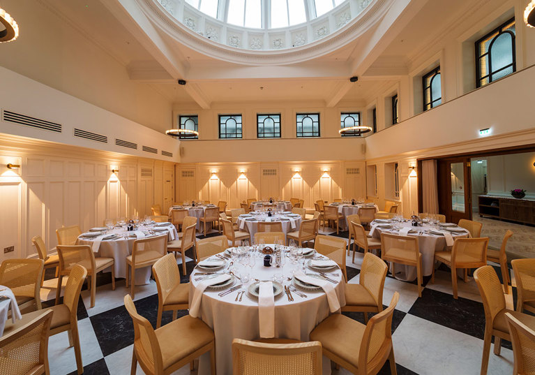 round tables with white linen and tableware set up for a wedding dinner below a glass dome on the ceiling