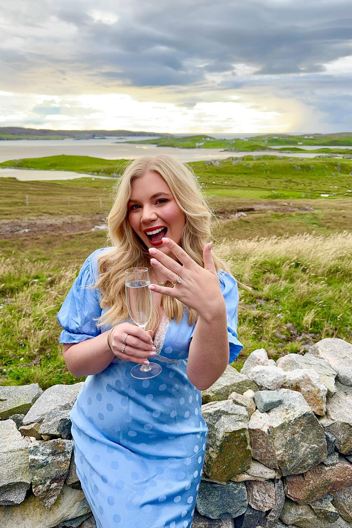 Fiancée in blue polka-dot dress smiling and showing her engagement ring while holding a Champagne flute with rugged coastline behind