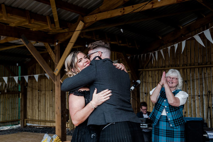 Wedding In A Shed With Bunting Hanging Featuring A Bride And Groom Embracing As A Person Wearing A Tartan Outfit Claps
