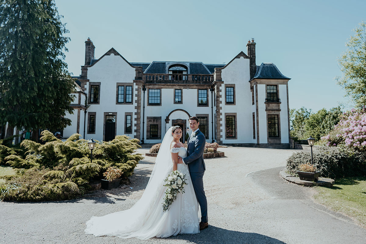 Bride and groom posing together in front of a grand white country house on a sunny day with blue sky overhead