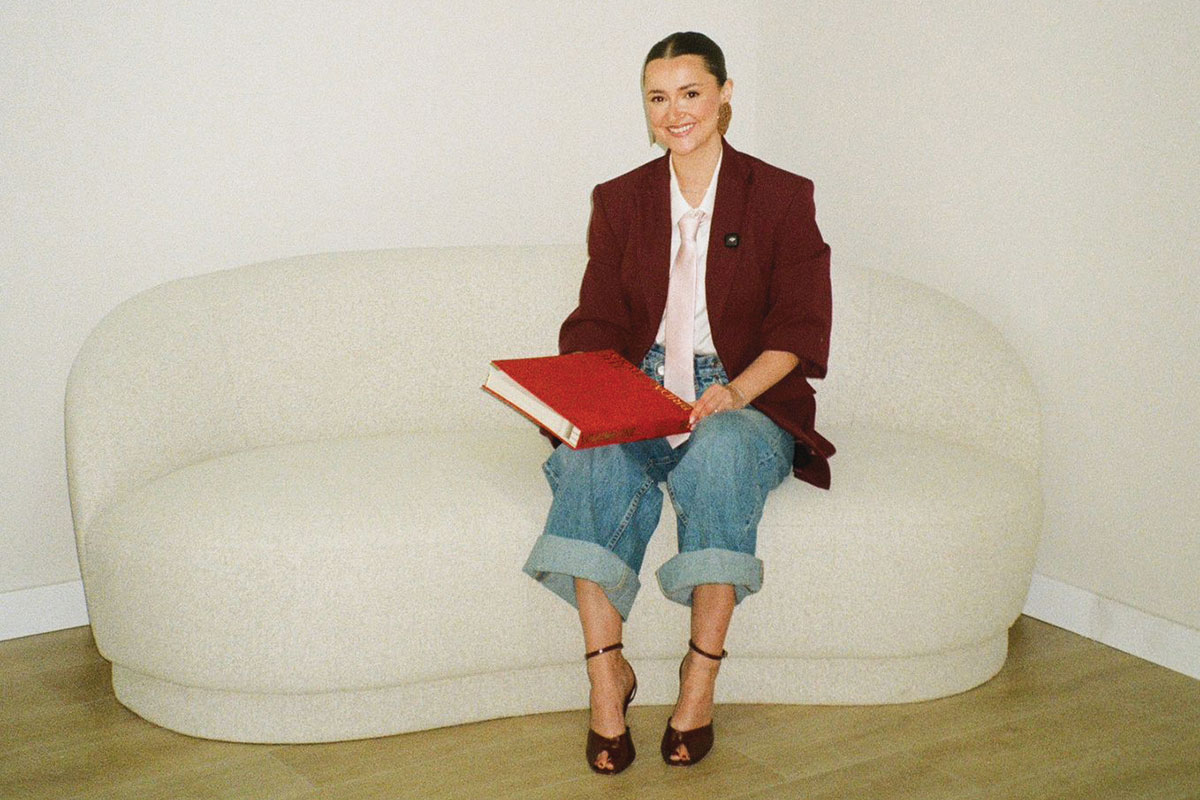 woman in burgundy blazer, jeans and pink silk tie sits on cream soda with a red book on her lap