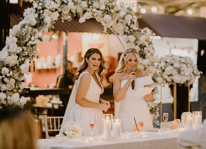 Two brides standing behind a long table that's set with candles in lanterns. There is a large white floral arch and white pillar floral arrangements in the background. One bride is holding a microphone and looking at a sheet of paper, while the second laughs