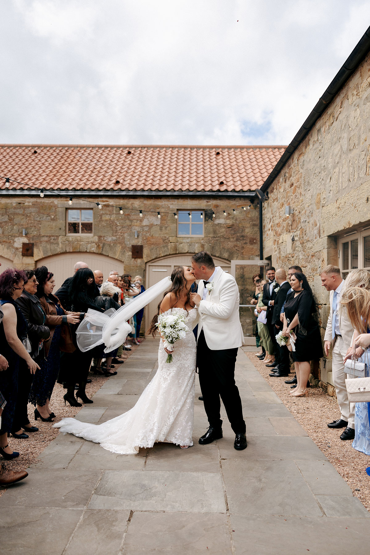Bride and groom share a kiss in the Falside Mill courtyard as guests cheer and the bride’s veil catches the wind.